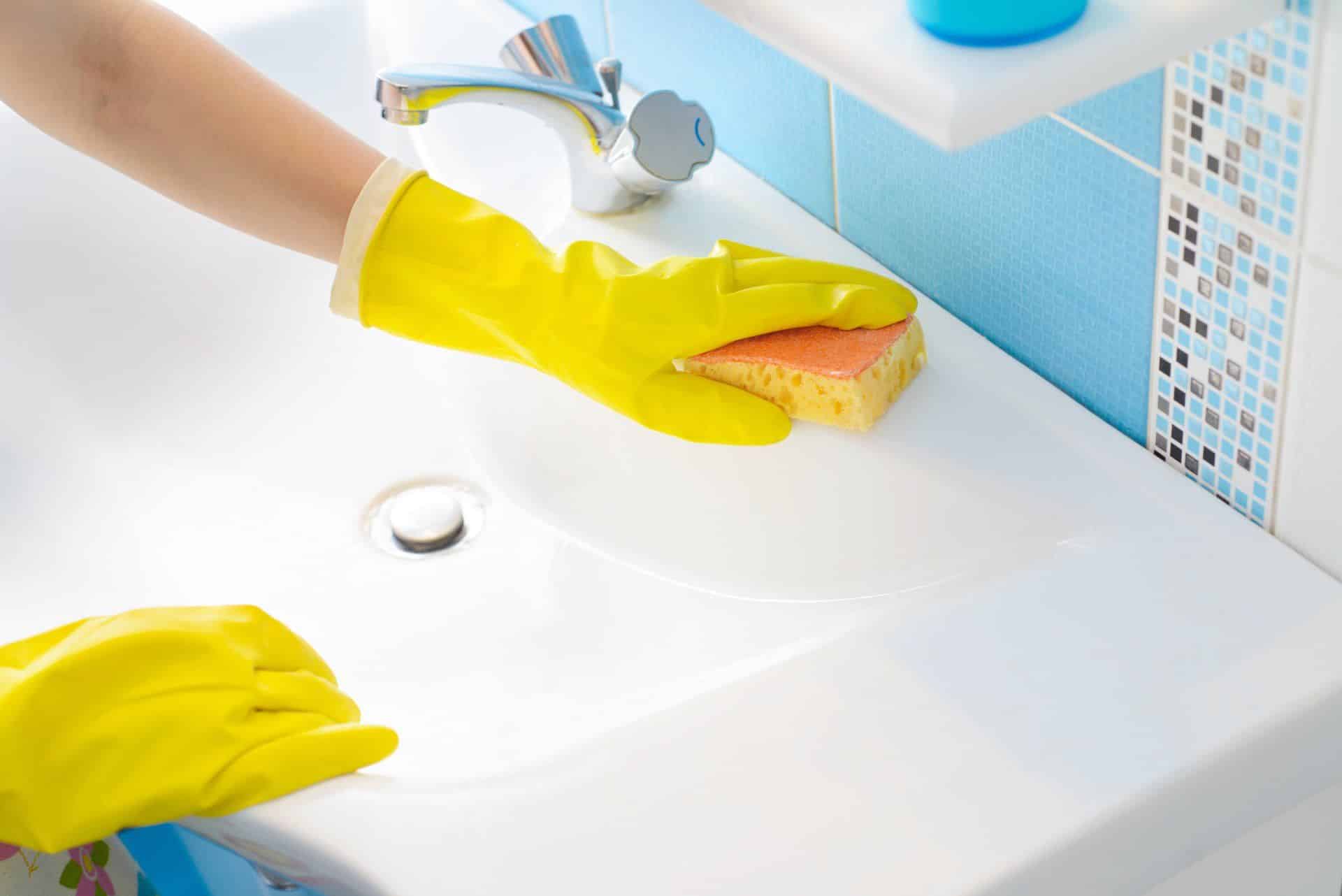 person cleaning bathroom sink with bleach and sponge while wearing yellow gloves