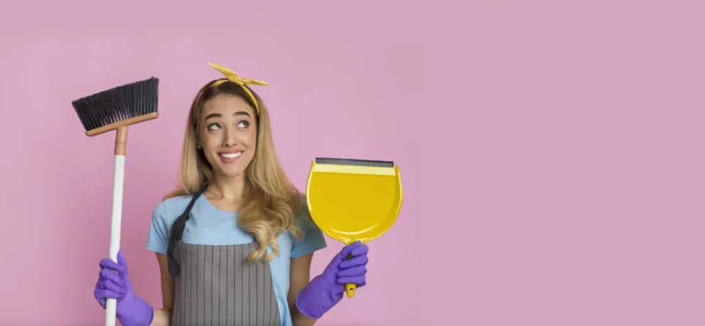 How to Clean Baseboards 6 A woman smiling and holding a broom and dustpan against a pink background