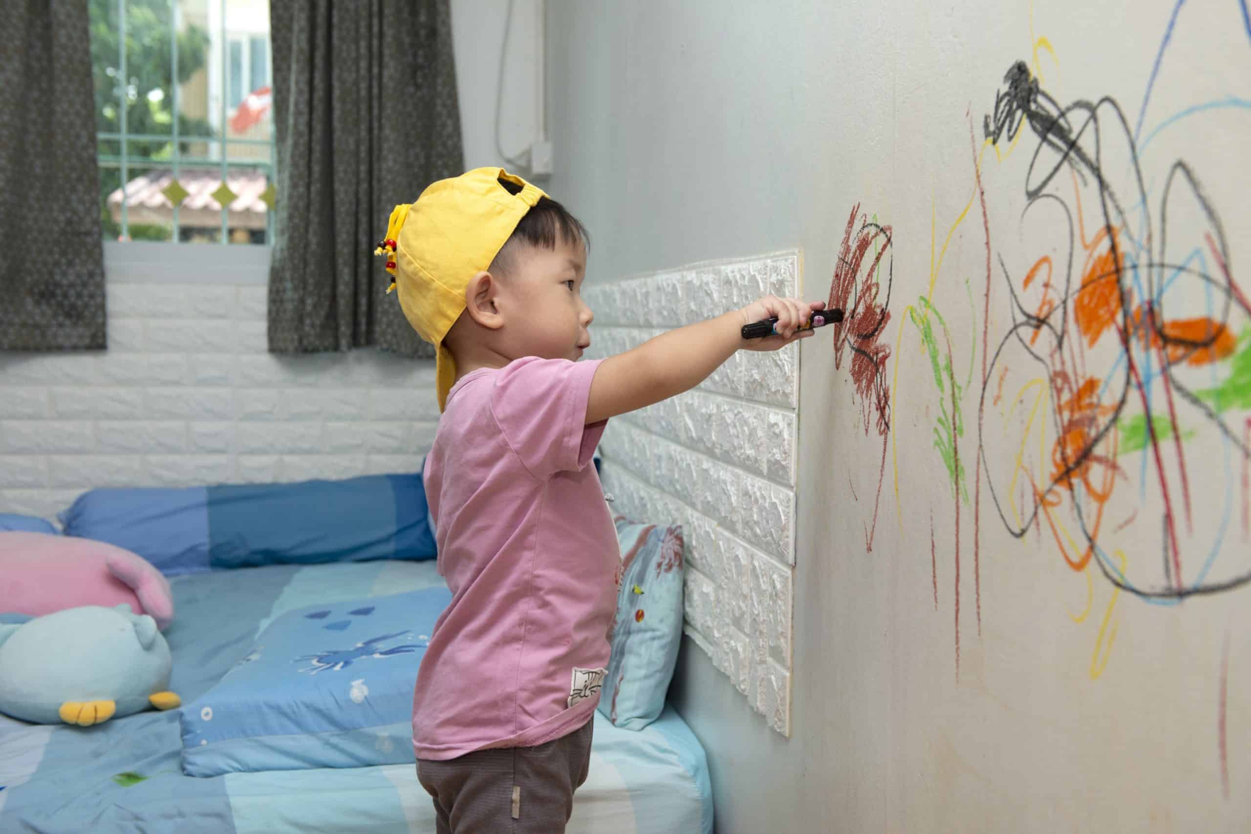A little boy using crayons to color on a wall.