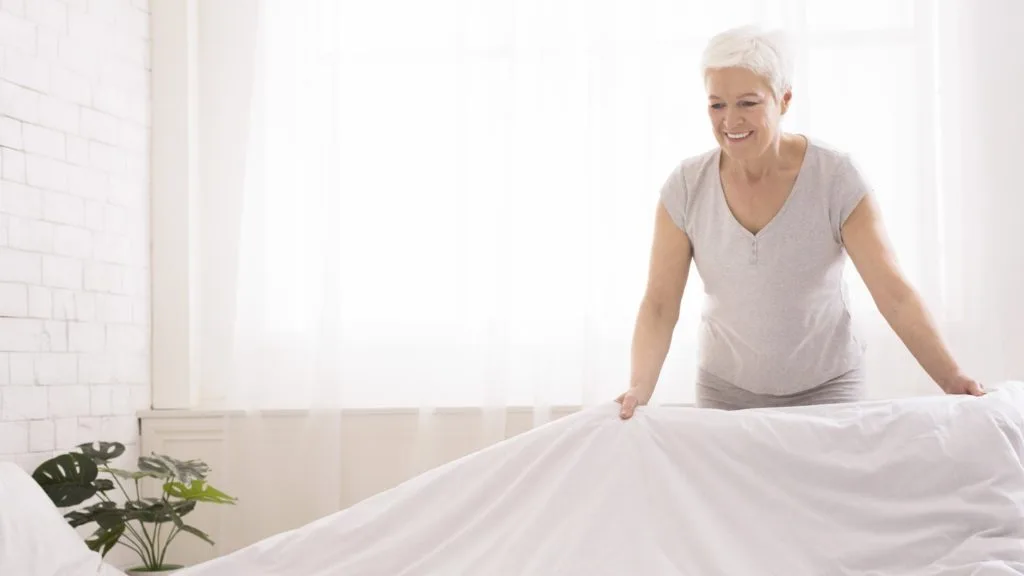 Learn How to Clean a Room in 5 Minutes 7 An elderly woman smiling and making her bed as as tep for cleaning a room in 5 minutes.