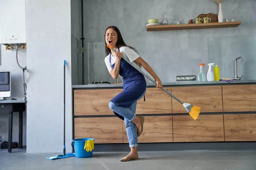 How to Clean Your House Cleaning Tools 9 A woman wearing an apron and singing into a broom while sweeping.