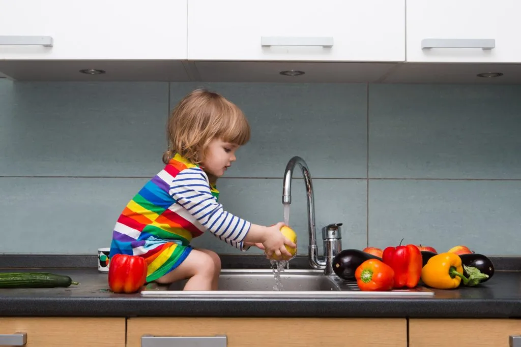 These 7 Chores for Kids Let You Delegate the Housework 2 A toddler sitting on a counter washing produce.