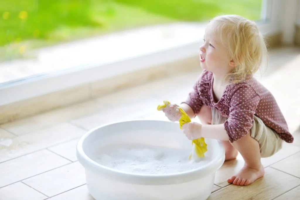 These 7 Chores for Kids Let You Delegate the Housework 4 A small blonde toddler using a cleaning bucket and gettign ready o clean baseboards.