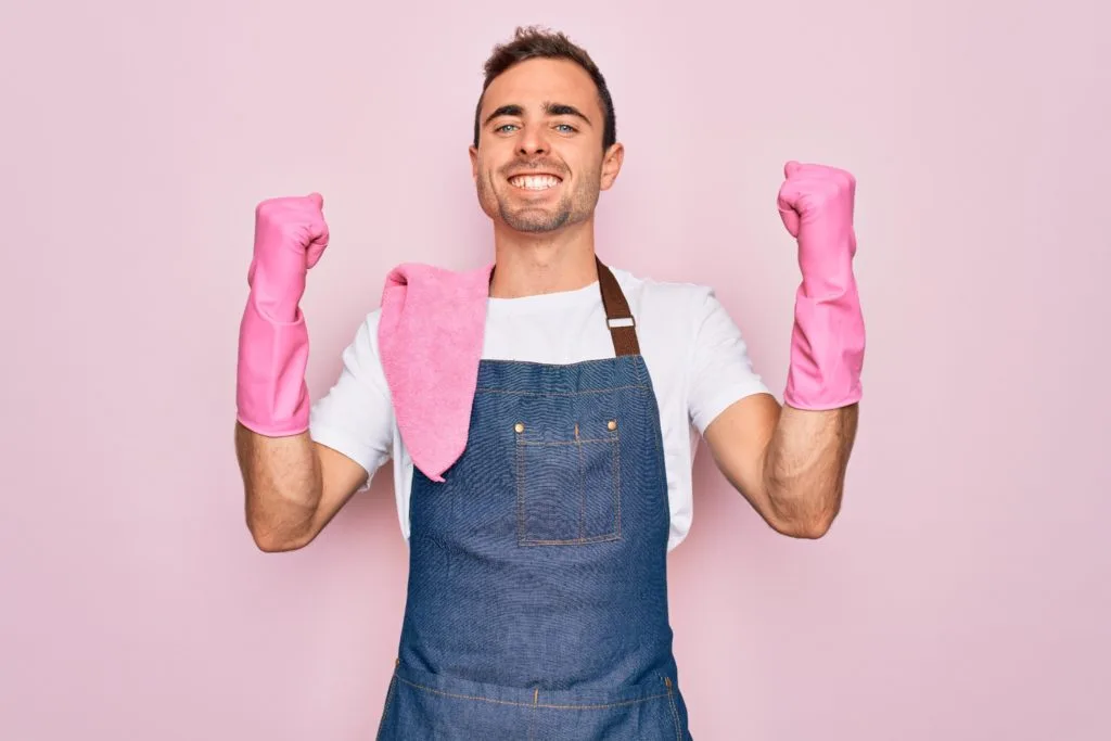 13 Things to Try and Make Cleaning Fun 1 A man wearing an apron and pink cleaning gloves flexing and smiling for a portrait.