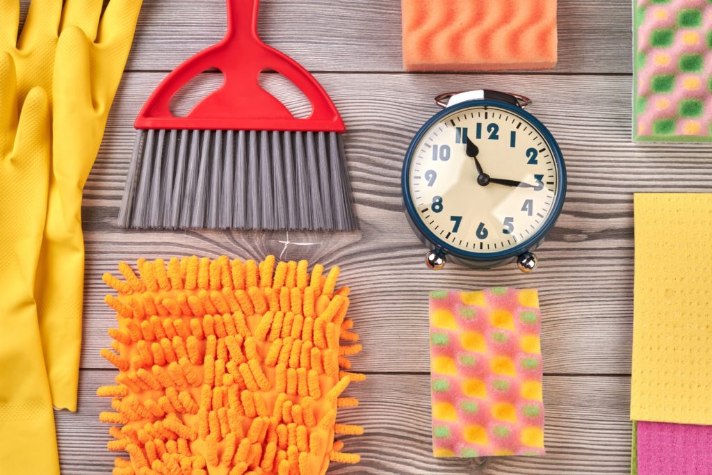 A flat lay of cleaning supplies and an alarm clock. Setting a timer is something your can try to make cleaning fun.