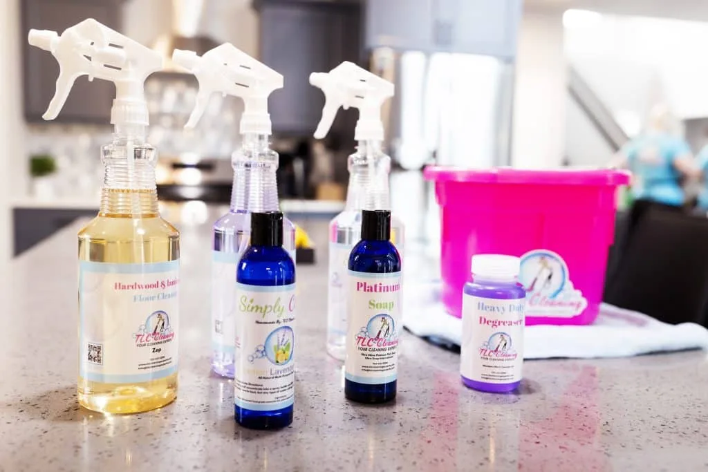 3 Cleaning Products to Avoid and 4 Products to Use Instead 2 A variety of labeled cleaning products are lined up on a speckled countertop, including sprays and a degreaser, with a pink cleaning bucket in the soft-focused background.