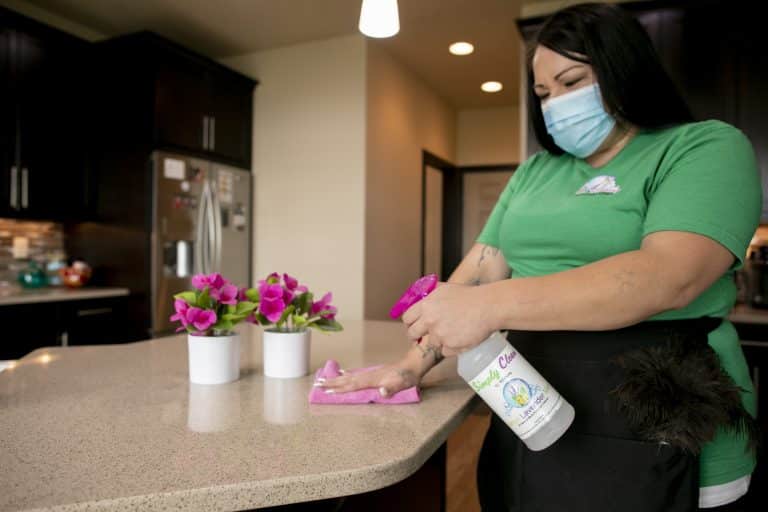 A person wearing a green shirt and a face mask uses cleaning products to wipe down a kitchen counter. Two small white pots with vibrant pink flowers sit on the countertop, adding a pop of color to the scene.
