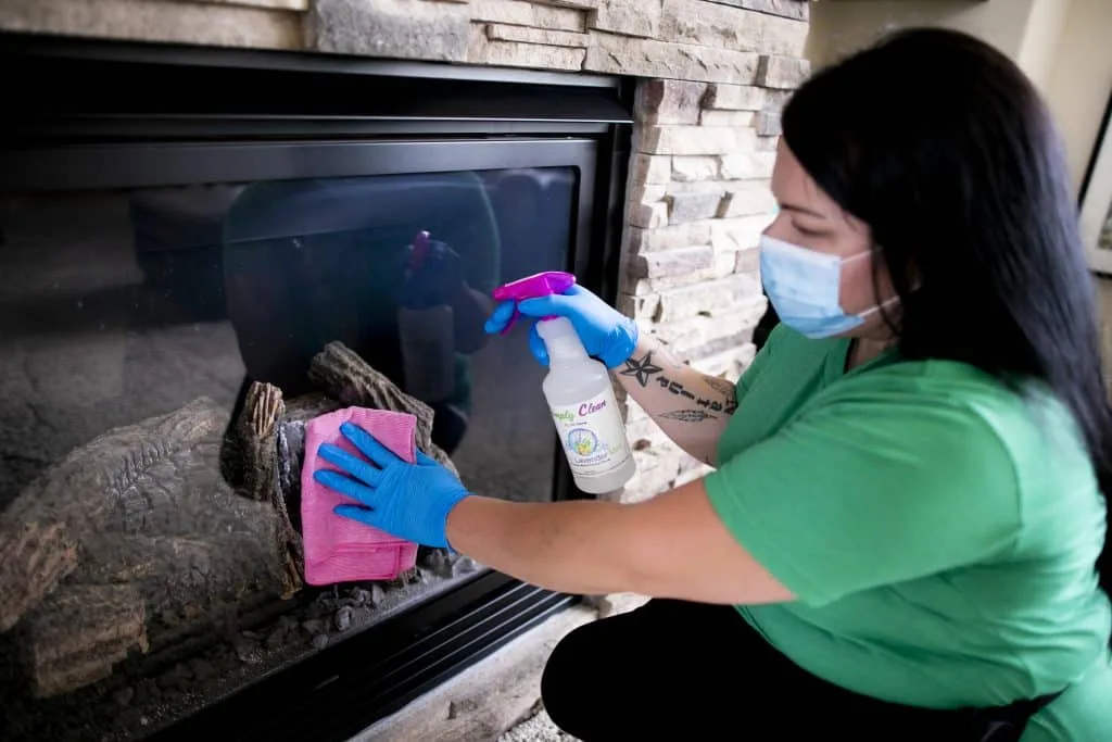 Tackling Hidden Dirt: The Power of Deep Cleaning Services 1 A person wearing a mask and blue gloves provides deep cleaning services, meticulously wiping down a fireplace glass door with a pink cloth and spray cleaner.