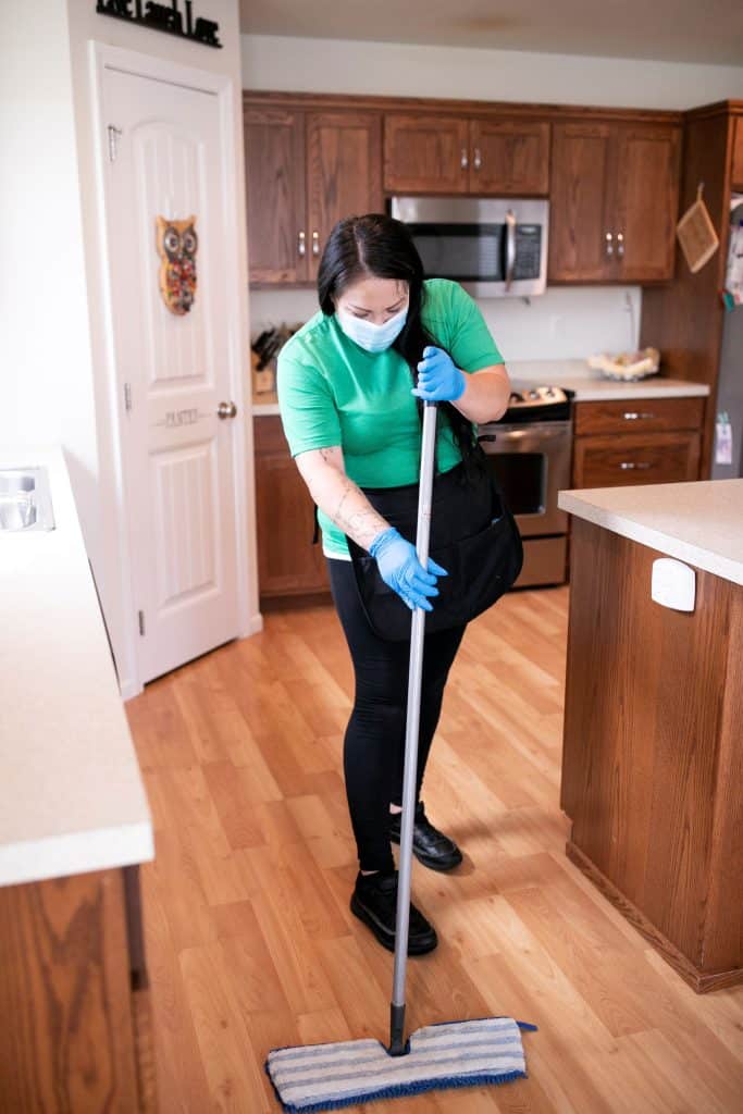 A professional cleaner in a green shirt and face mask performs move-out cleaning in a modern kitchen, ensuring thorough cleanliness.