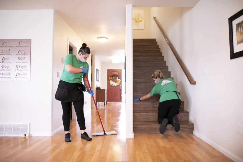 Move-In Cleaning: How to Start Fresh with a Spotless Home 2 Two cleaning professionals in green shirts performing a thorough cleaning: one mopping the hardwood floor, and the other scrubbing the stairs in a well-lit home entryway.