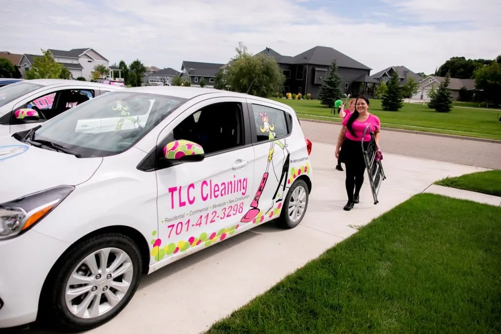 Move-Out Cleaning Woes: Ensure a Stress-Free Deposit Return 2 A TLC Cleaning service vehicle is parked in a residential neighborhood, with a professional cleaner in a pink shirt walking towards a house with cleaning supplies.