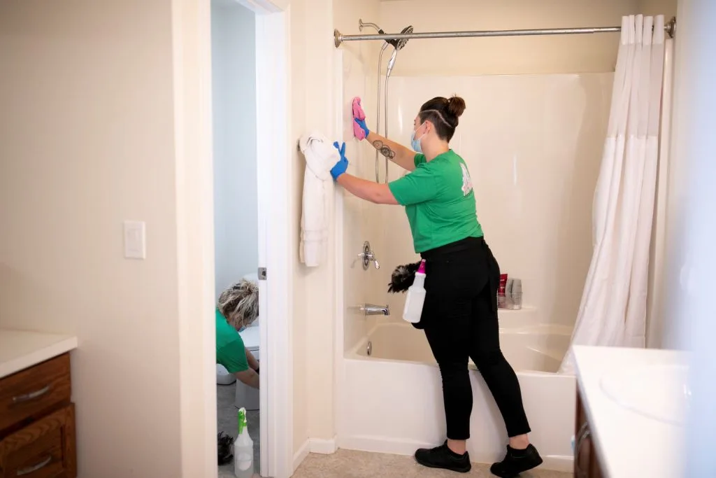 Top Bathroom Cleaning Tips for a Sparkling Fresh Lavatory 4 Two professional cleaners in green uniforms clean a bathroom, with one scrubbing the shower wall and the other cleaning the toilet in the adjacent room.