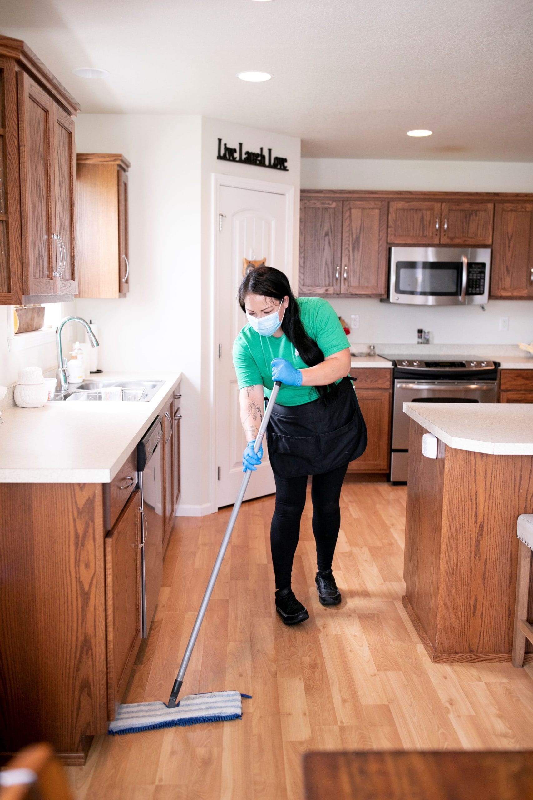 A professional cleaner wearing gloves and a mask mops the floor of a modern kitchen, ensuring a thorough cleaning of the space.